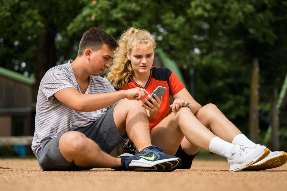 Zwei Jugendliche sitzen auf dem Boden und betrachten gemeinsam ein Smartphone. Ein Mädchen trägt ein rotes sportliches Oberteil.